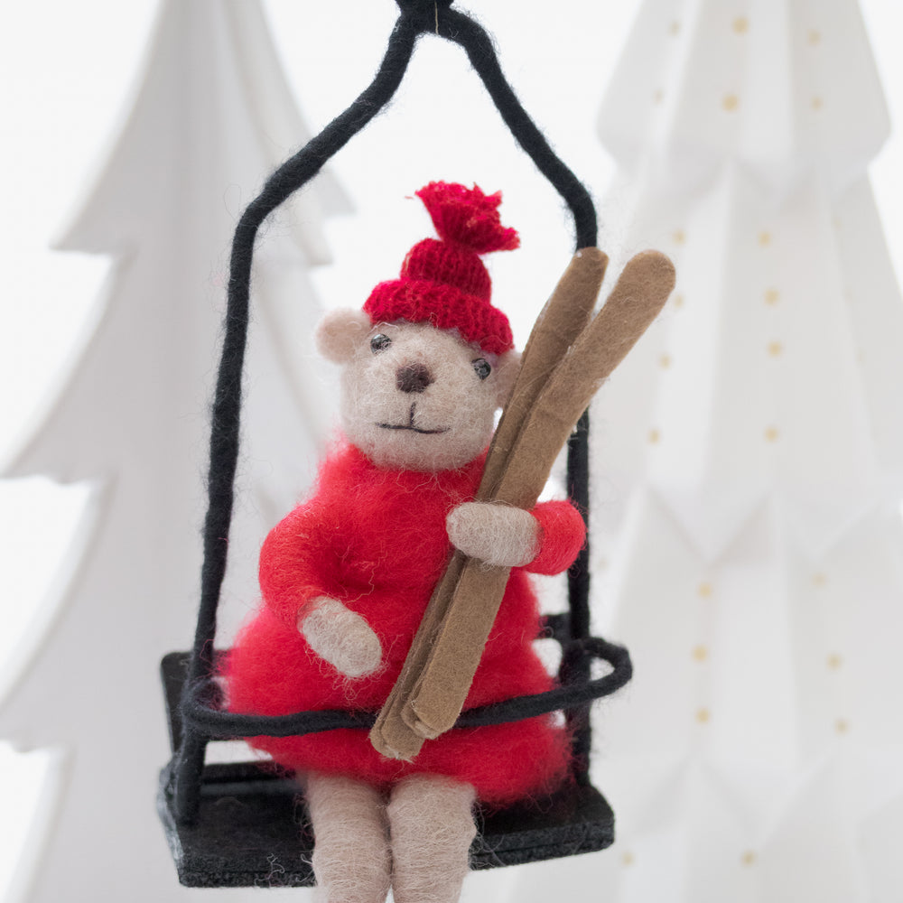 Decorative teddy bear ornament in a red outfit and hat, holding skis, on a black ski lift .